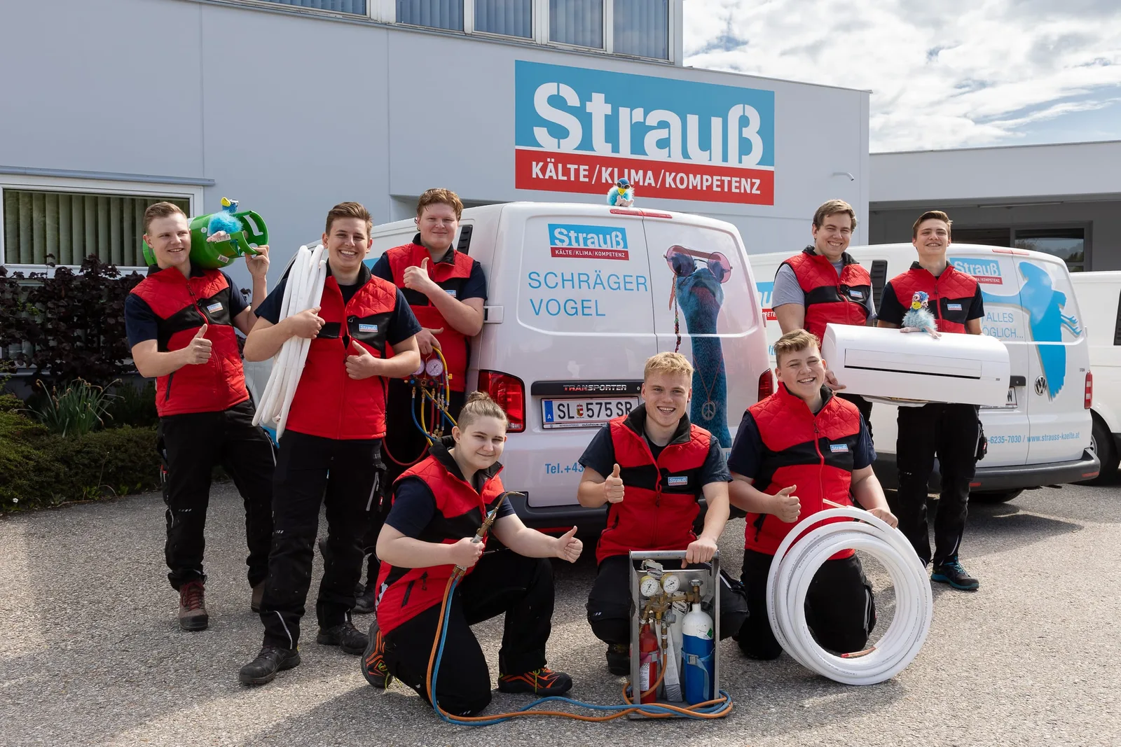 Strauß-Lehrlinge in rot-schwarzer Arbeitskleidung mit Rohren und Werkzeug vor einem Firmenwagen