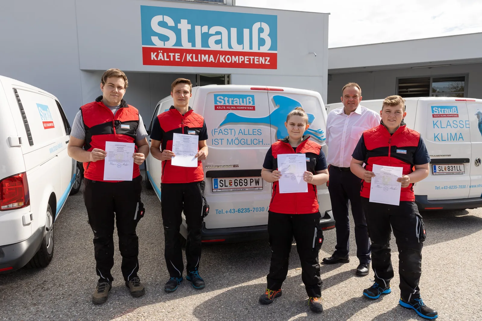 Vier Lehrlinge in Arbeitskleidung mit ihren Zeugnissen vor den Firmenwagen, daneben ein Mann im weißen Hemd
