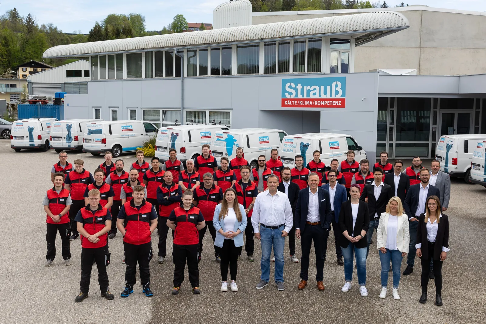 Das ganze Strauß-Team in Arbeitskleidung vor dem Firmensitz mit Schild und Firmenwagen